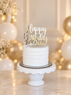 A white frosted cake on a white pedestal stand with a gold 'Happy Birthday Dr. Niki' acrylic topper, set against a warm, softly blurred background with balloons and fairy lights.