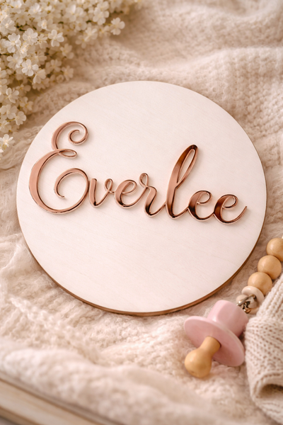 Round wooden sign with 'Everlee' engraved, placed on a soft fabric background with baby items.