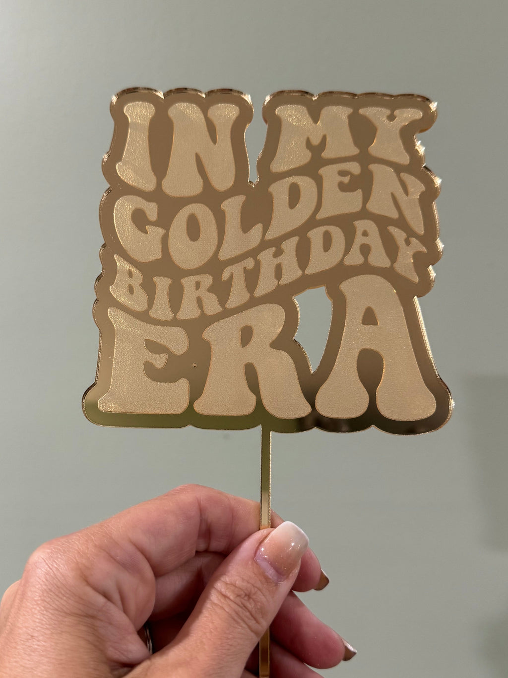 In My Golden Birthday Era gold mirrored acrylic cake topper held by hand to show its size and reflective finish