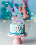 A personalized birthday cake with a mermaid-themed topper, featuring glitter and multiple colors, placed on a stand with a decorative background.
