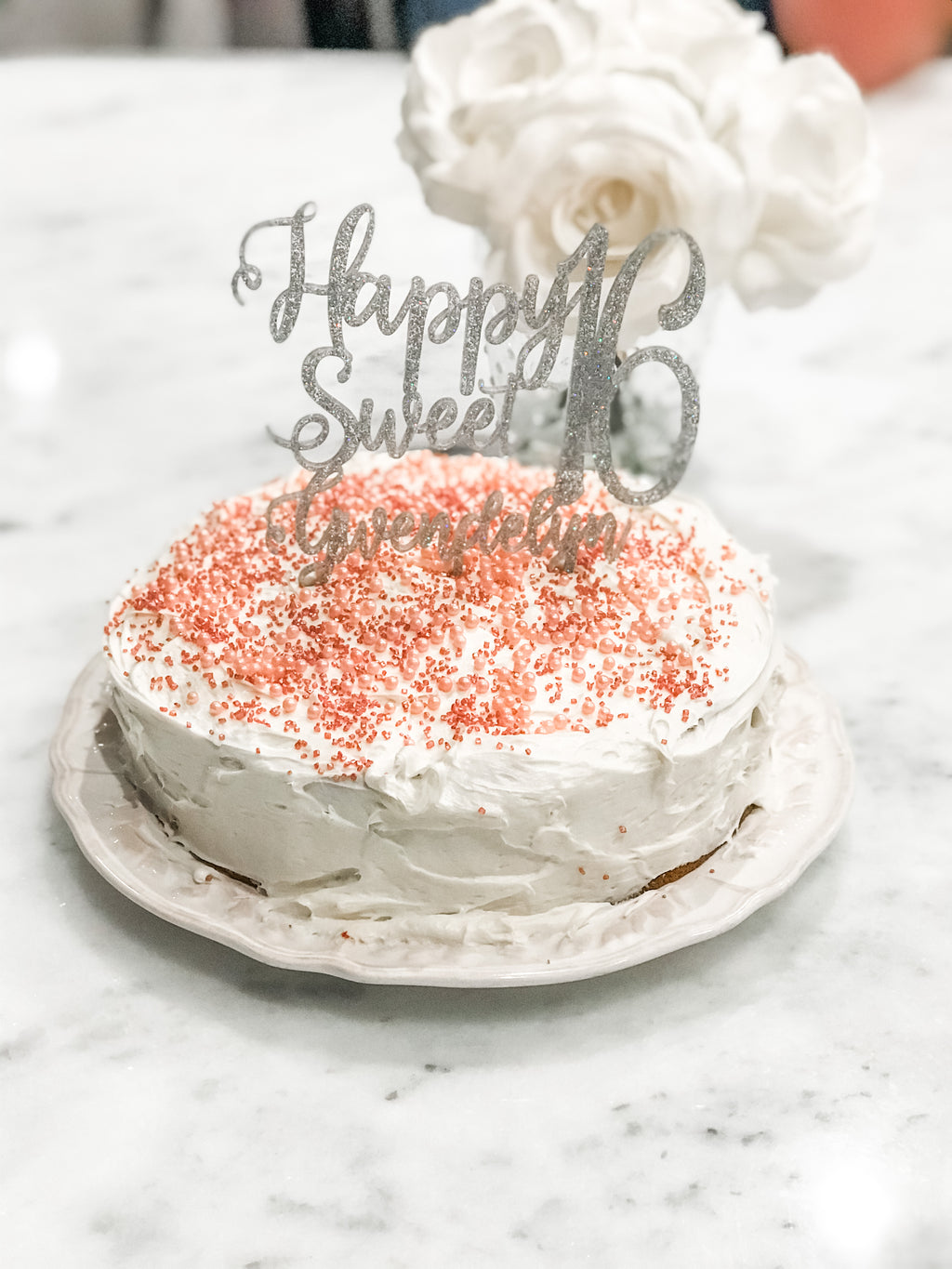 A frosted birthday cake topped with orange sprinkles and a silver glitter 'Happy Sweet 16' topper, sitting on a white plate on a marble surface with white flowers in the background.