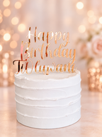 A white frosted cake topped with a rose gold acrylic 'Happy Birthday To Uluwanti' script cake topper, set against a warm, blurred bokeh background with soft lights and flowers.