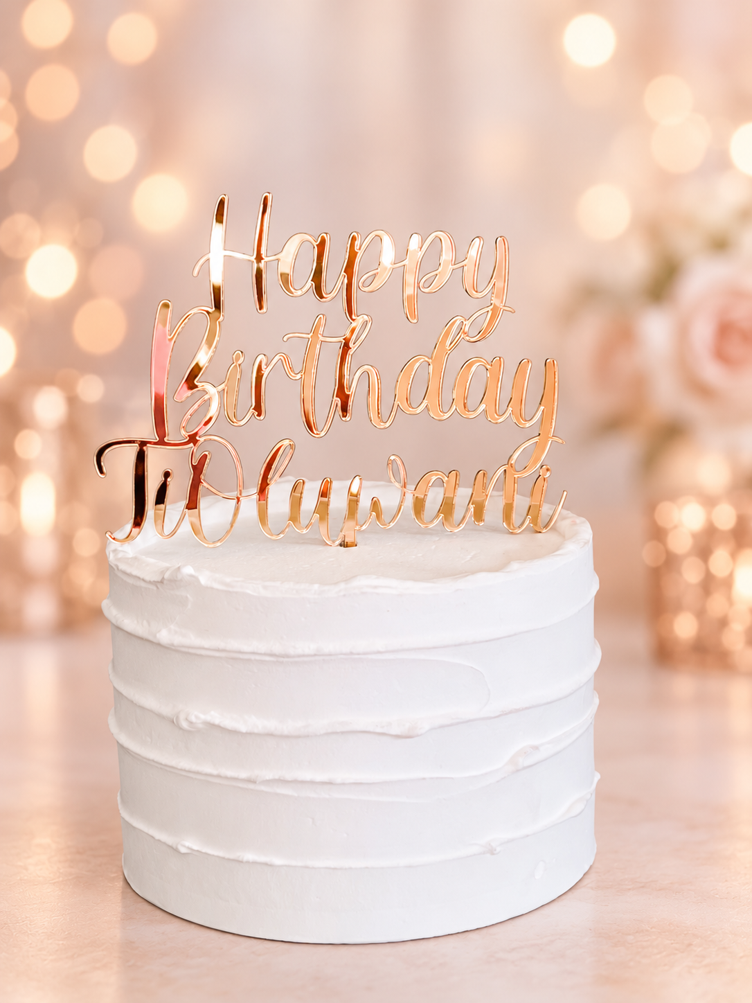 A white frosted cake topped with a rose gold acrylic 'Happy Birthday To Uluwanti' script cake topper, set against a warm, blurred bokeh background with soft lights and flowers.