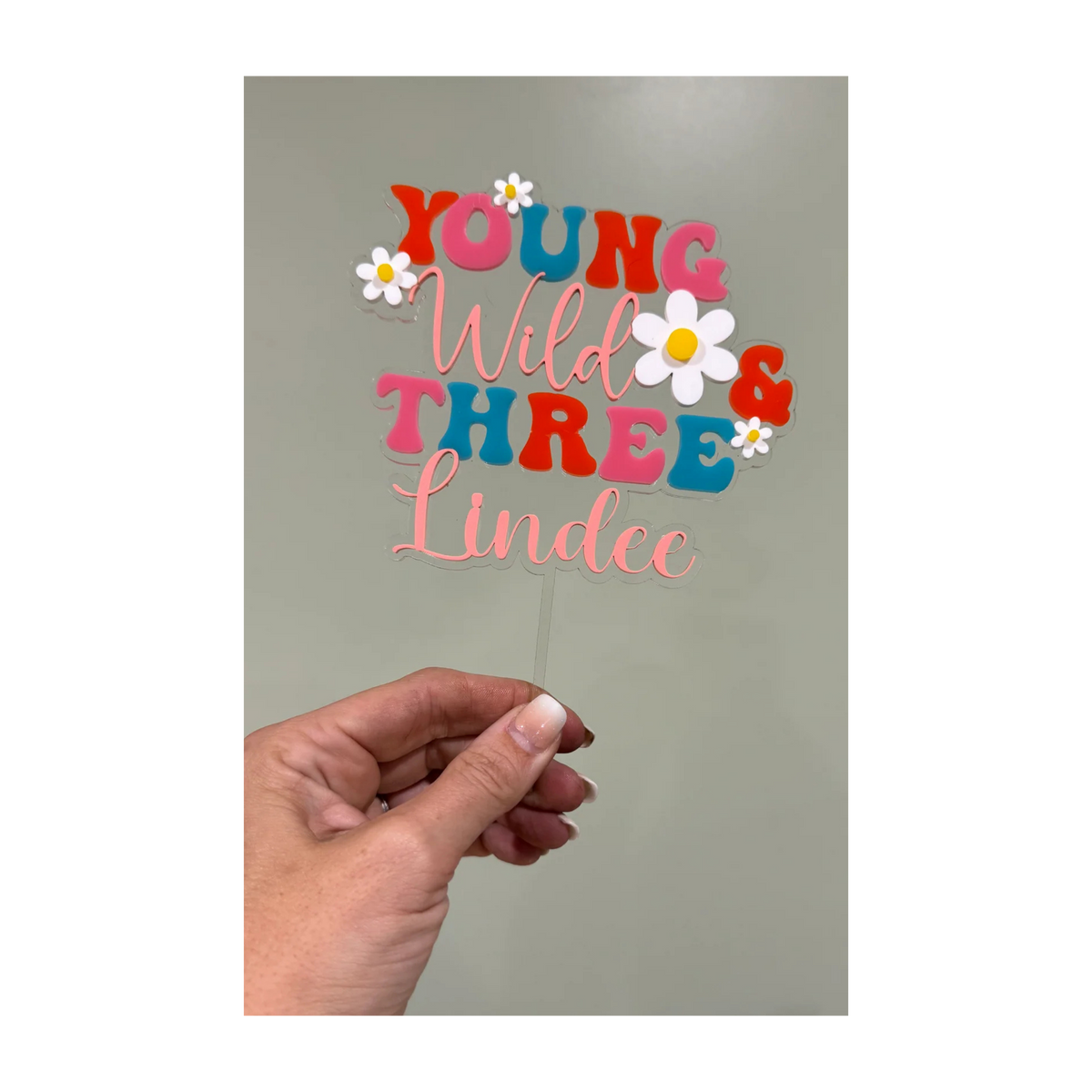 Hand holding a personalized Young Wild and Three layered acrylic cake topper with the name Lindee
